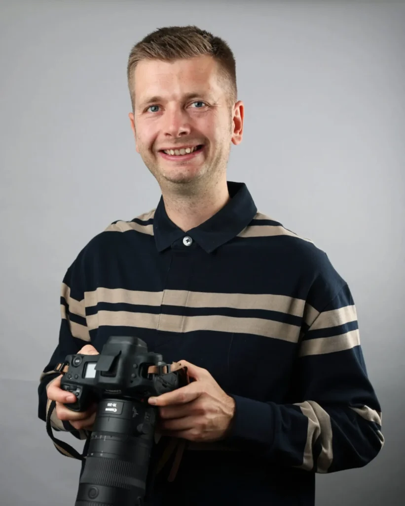 Portrait of Nathan Stirk holding a camera, smiling against a grey background.