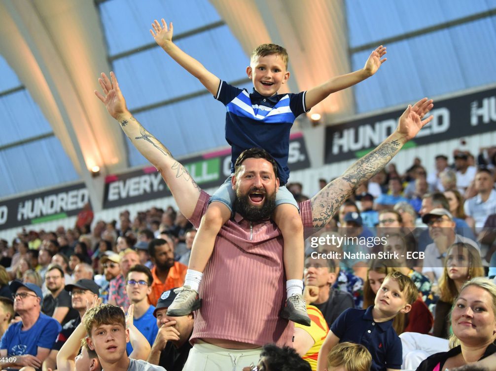 A father joyfully lifts his son on his shoulders during a sports event, surrounded by a cheering crowd.