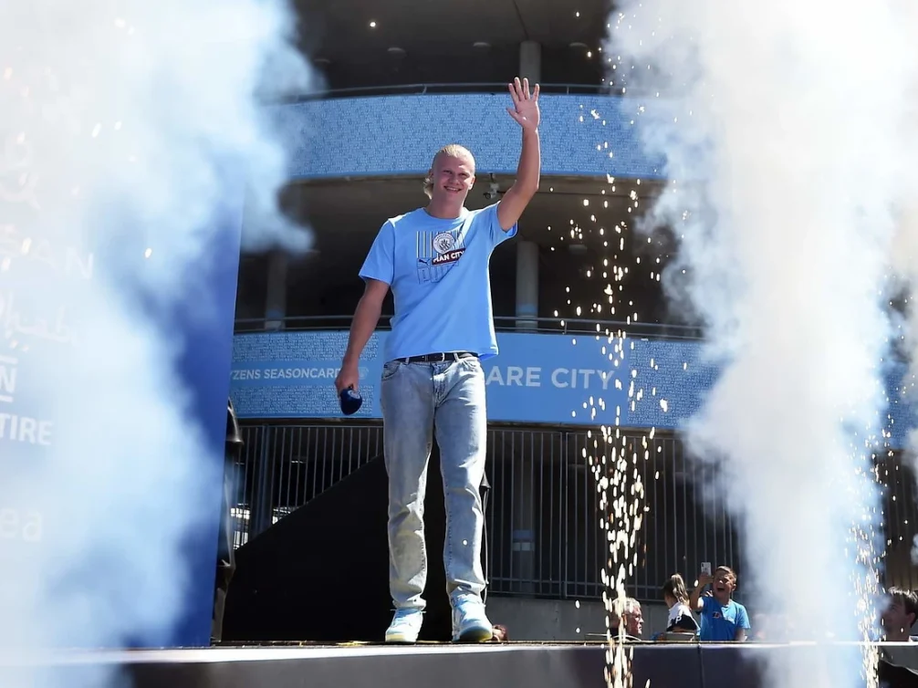 A footballer in a Manchester City shirt greets the crowd at an event.