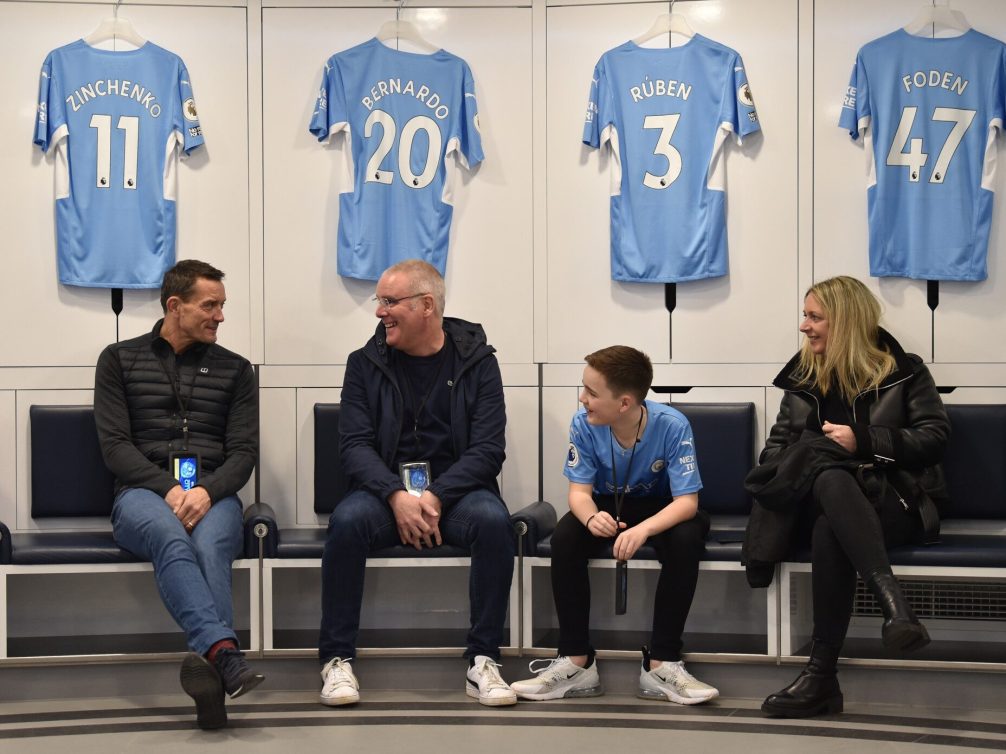 Inside the Manchester City locker room featuring jerseys and smiling fans.